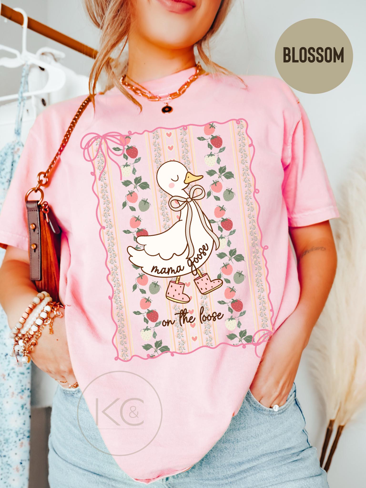 May include: Pink t-shirt with a graphic of a cartoon goose wearing boots and a bow. The text "mama goose on the loose" is printed below the goose. The shirt has a floral and strawberry design around the graphic.