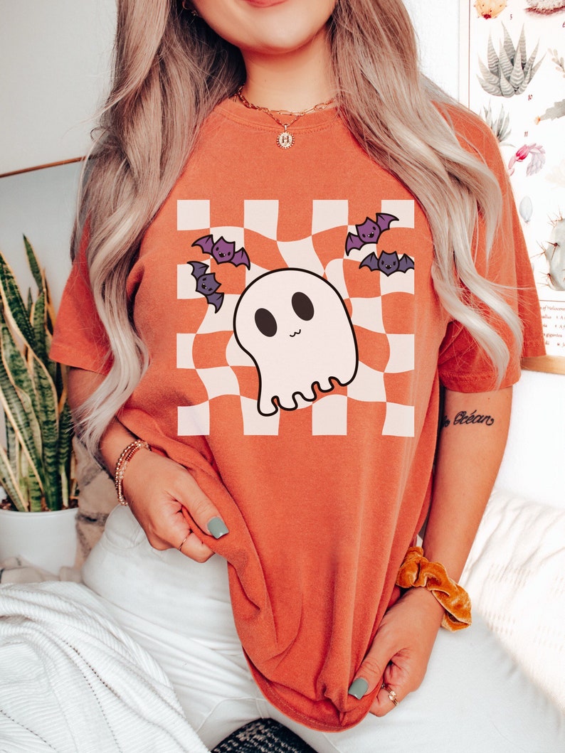 May include: Orange t-shirt with a white and black checkered pattern featuring a cartoon ghost and bats. The text "Boo" is written on the sleeve.