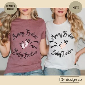 Może przedstawiać: Dwie koszulki z napisem "Mommy Besties" i "Baby Besties" w kółku wokół pary różowych śladów stóp dziecka. Jedna koszulka jest w kolorze heathered mauve, a druga biała.