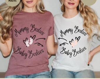 May include: Two t-shirts with the text "Mommy Besties" and "Baby Besties" in a circle around a pair of pink baby footprints. One shirt is heather mauve and the other is white.