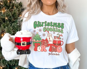 May include: White t-shirt with a Christmas-themed graphic. The design includes the text "Christmas Cookies For Two please" in green and red, along with illustrations of cookies, a Christmas tree, and a mug of hot chocolate. A red Santa mug is held in the image.