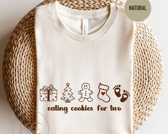 May include: Cream-colored t-shirt with a brown graphic of a gift, Christmas tree, gingerbread man, stocking, and footprints. The text "eating cookies for two" is below the graphic. The shirt is on a woven surface.