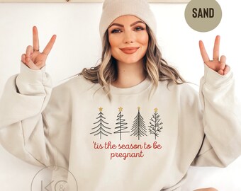 May include: Beige sweatshirt with a Christmas tree design and the text "'tis the season to be pregnant". The sweatshirt also features a pair of golden footprints. The text "For oversized look order 1-2 sizes up" is at the top.