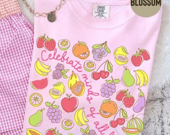 May include: A light pink t-shirt featuring a colorful graphic print of various fruits and vegetables, including strawberries, bananas, grapes, and oranges. The text "Celebrate minds of all kinds" is printed in a cursive font across the center.