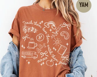 May include: A rust-colored t-shirt featuring white line art graphics including a school bus, an apple, a coffee mug, a rainbow, a pencil, a notebook, and the text "Teach them, love them, watch them grow" with musical notes and bows.
