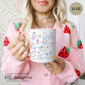 May include: White ceramic mug with a pink rim and handle. The mug features a colorful illustration of cats playing with yarn and knitting needles. The text "11 oz" is printed on the mug.