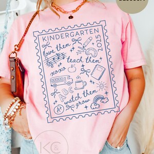 May include: Pink t-shirt with a blue graphic design featuring the word "KINDERGARTEN" and the phrases "love them, teach them, watch them grow." The design includes school-related illustrations such as a pencil, apple, and school bus. The word "BLOSSOM" is in a circle.
