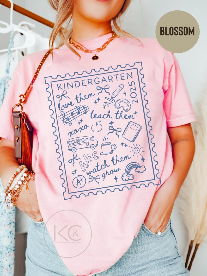 May include: Pink t-shirt with a blue graphic design featuring the word "KINDERGARTEN" and the phrases "love them, teach them, watch them grow." The design includes school-related illustrations such as a pencil, apple, and school bus. The word "BLOSSOM" is in a circle.