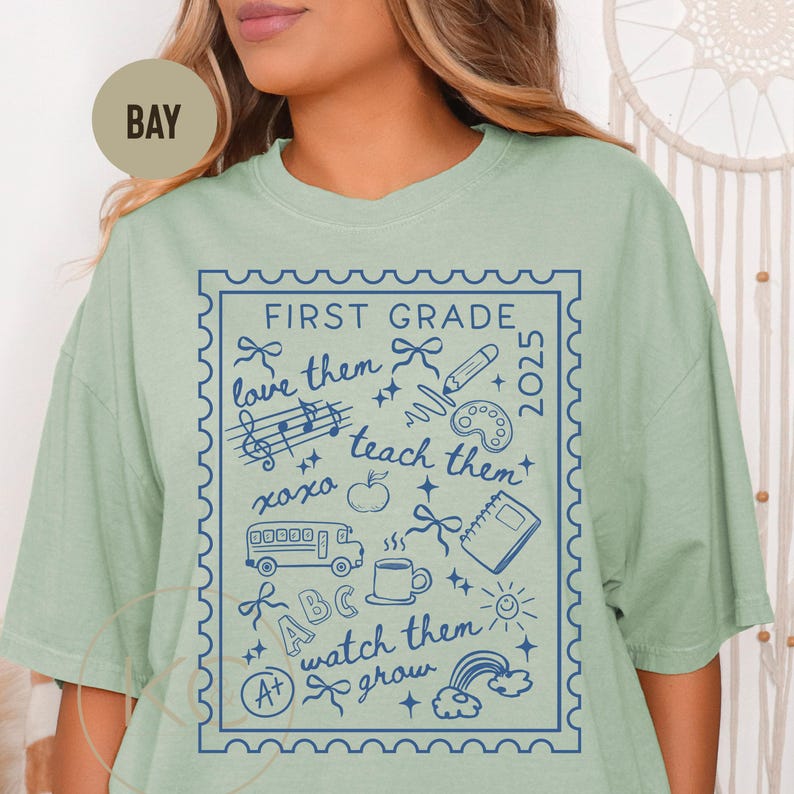 May include: Pale green t-shirt featuring a blue graphic. The design, within a stamp-like border, says "FIRST GRADE" and "2025", with school-related illustrations like a bus, apple, and rainbow.