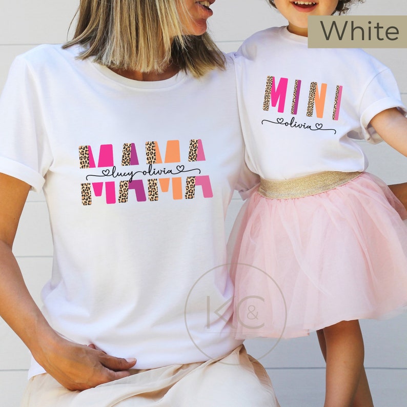 May include: White t-shirts with the words "MAMA" and "MINI" in pink, orange, and purple with a leopard print design. The text "lucy olivia" is written in black below the word "MAMA" and the text "olivia" is written in black below the word "MINI".
