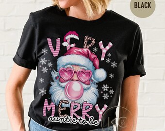 May include: Black t-shirt with a graphic of Santa Claus wearing heart-shaped sunglasses and blowing a bubblegum bubble. The text reads "VERY MERRY auntie to be" in pink and white lettering, with snowflake accents.