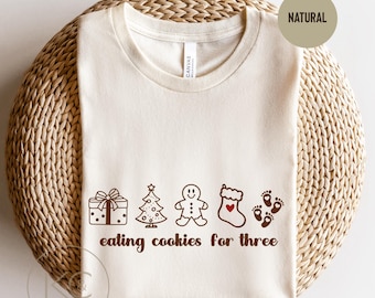May include: Cream-colored t-shirt with a brown graphic of a gift, Christmas tree, gingerbread man, stocking, and footprints. The text "eating cookies for three" is below the graphics. The shirt is folded on a woven surface.