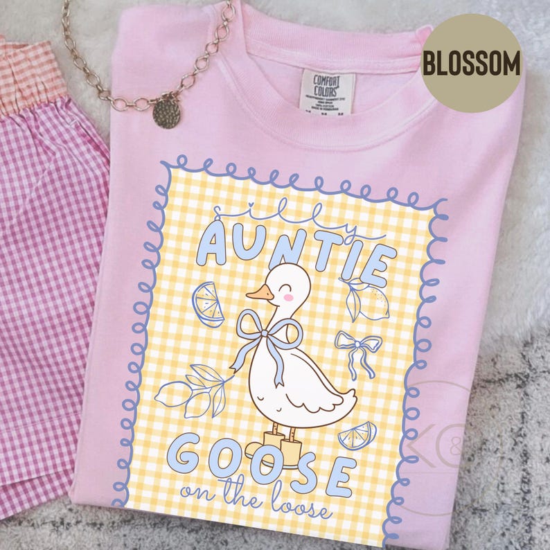 May include: A light pink t-shirt with a graphic featuring a cartoon goose wearing a blue bow and boots, with the words "Auntie Goose on the Loose". The design is set against a yellow and white checkered background.