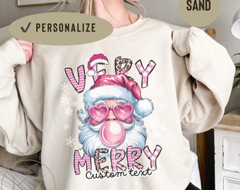 May include: Sand-colored sweatshirt featuring a Santa Claus graphic with heart-shaped sunglasses blowing a bubblegum bubble. The text "VERY MERRY" is displayed in pink and black, with space for custom text.