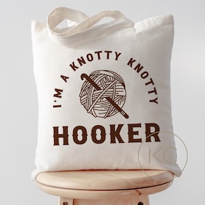 May include: A white tote bag with brown text that reads "I'm a knotty knotty hooker" with a drawing of a ball of yarn and a crochet hook.