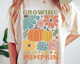 May include: Ivory-colored t-shirt with a colorful floral and pumpkin design. The text reads "GROWING A LITTLE PUMPKIN." Additional text advises ordering 1-2 sizes up for an oversized look. The design includes orange, yellow, blue, and pink flowers.