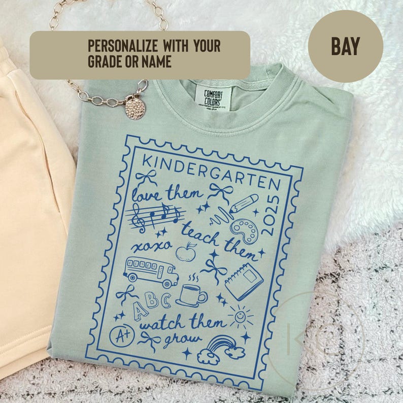 May include: A light green Comfort Colors t-shirt with a blue Kindergarten design. The design includes the text "Kindergarten 2025", musical notes, a school bus, and other school-related icons. The shirt also has the text "love them", "teach them", and "watch them grow".