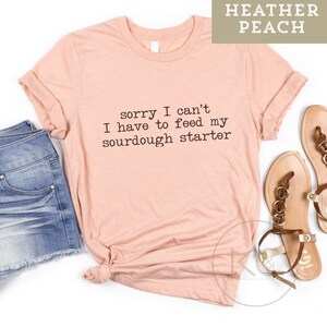 May include: A peach colored t-shirt with the text "sorry I can't I have to feed my sourdough starter" printed on the front.