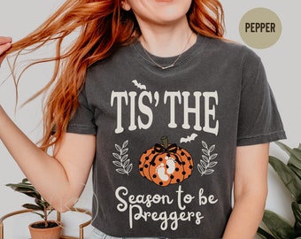 May include: Dark gray t-shirt with the text "Tis' the Season to be Preggers" in white script. The shirt features a pumpkin graphic with baby footprints and bats, perfect for Halloween or a pregnancy announcement.