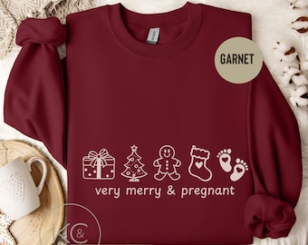 May include: Garnet-colored sweatshirt with white graphic of Christmas icons and text "very merry & pregnant." Features a gift, tree, gingerbread man, stocking, and footprints. The sweatshirt is displayed on a white fur-like surface.