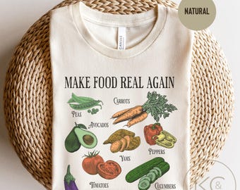 May include: A beige t-shirt with the text "Make Food Real Again" printed on the front. The shirt features a graphic of various vegetables and fruits, including peas, avocados, carrots, yams, peppers, cucumbers, and tomatoes.
