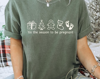 May include: A moss green t-shirt with a white graphic that includes a gift, Christmas tree, gingerbread man, stocking, and footprints. The text "'tis the season to be pregnant" is below the graphic.