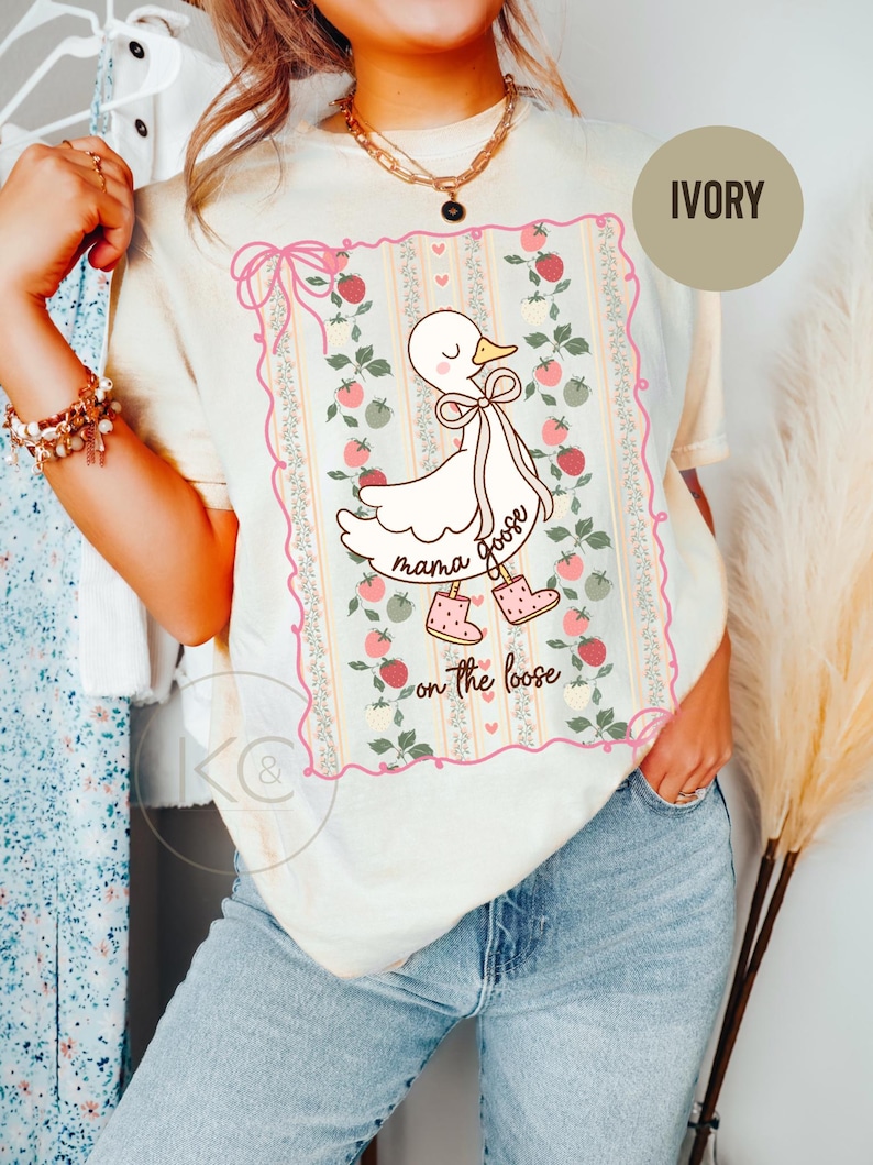 May include: Ivory-colored t-shirt featuring a cartoon goose with a bow, boots, and the words "mama goose on the loose." The design is framed by a pink floral border. The shirt is a light, neutral color.