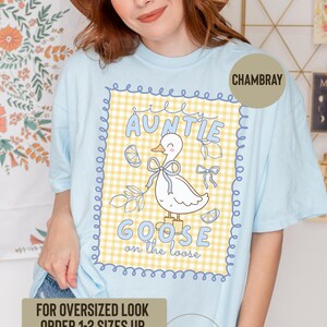 May include: Light blue chambray t-shirt with a graphic featuring a cartoon goose wearing a bow, surrounded by lemons and the text "AUNTIE GOOSE on the loose." The shirt also has the text "FOR OVERSIZED LOOK ORDER 1-2 SIZES UP."