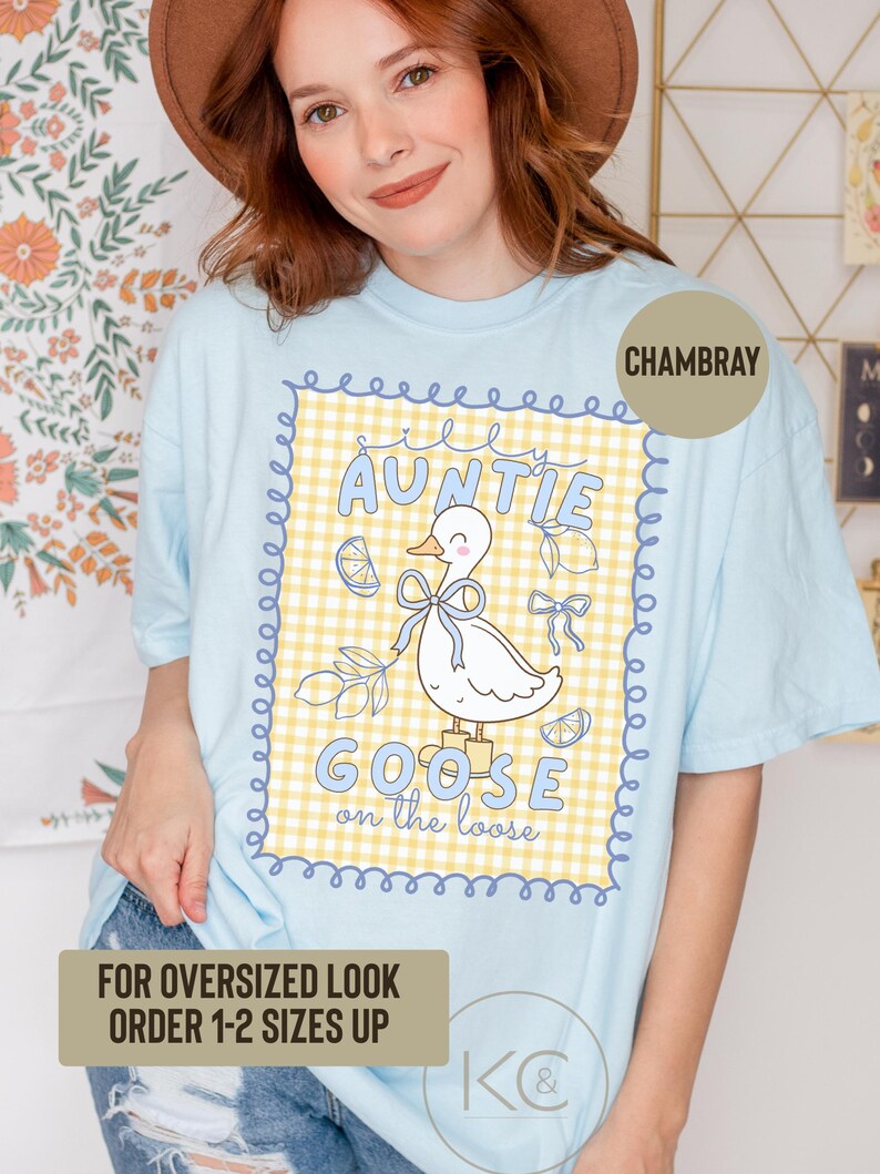 May include: Light blue chambray t-shirt with a graphic featuring a cartoon goose wearing a bow, surrounded by lemons and the text "AUNTIE GOOSE on the loose." The shirt also has the text "FOR OVERSIZED LOOK ORDER 1-2 SIZES UP."