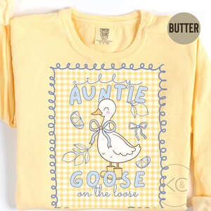 May include: A yellow sweatshirt with a graphic featuring a white goose wearing boots and a blue bow. The text reads "Silly Auntie Goose on the Loose" in blue lettering, set against a yellow and white gingham pattern.