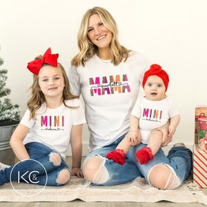 May include: Three white t-shirts with colorful text. The largest shirt says "Mama Scarlett" with a pink heart. The medium shirt says "Mini Olivia" with a pink heart. The smallest shirt says "Mini Olivia" with a pink heart.