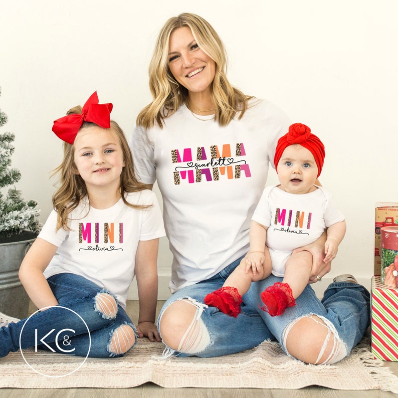 May include: Three white t-shirts with colorful text. The largest shirt says "Mama Scarlett" with a pink heart. The medium shirt says "Mini Olivia" with a pink heart. The smallest shirt says "Mini Olivia" with a pink heart.