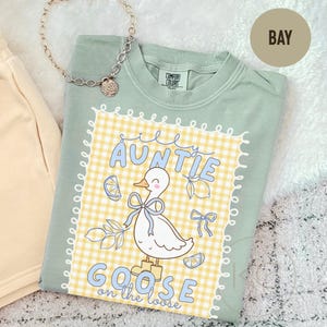May include: A sage green Comfort Colors t-shirt with a graphic that reads "Auntie Goose on the Loose." The graphic features a cartoon goose with a blue bow, lemons, and a yellow gingham background. A silver necklace is also visible.