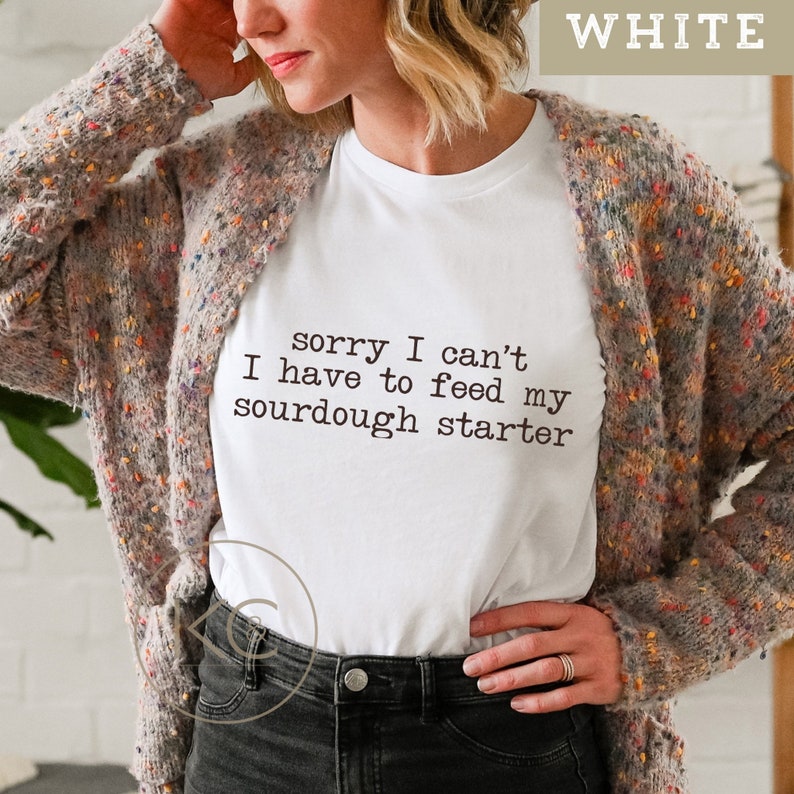 May include: White t-shirt with the text "sorry I can't I have to feed my sourdough starter" printed on it.
