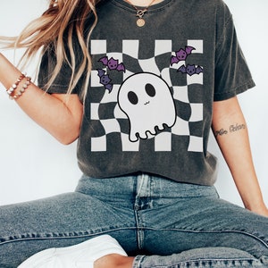 May include: A dark grey t-shirt with a white checkered pattern and a cartoon ghost with bats flying around it. The shirt has a relaxed fit and a crew neck.