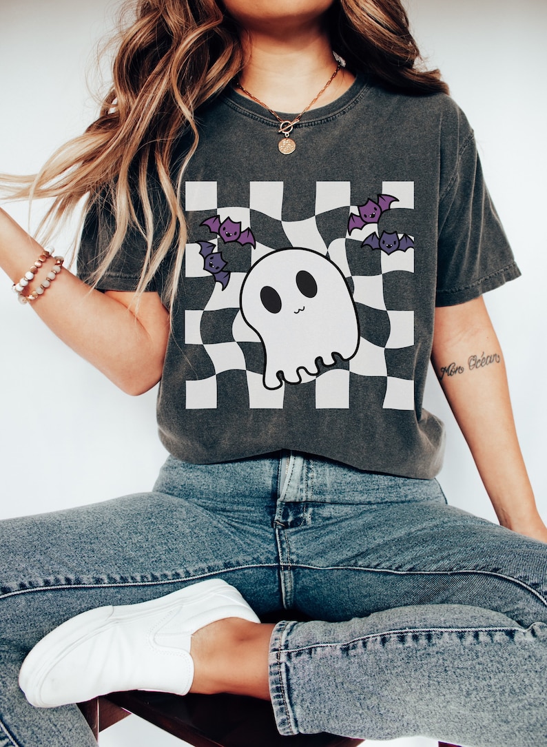 May include: A dark grey t-shirt with a white checkered pattern and a cartoon ghost with bats flying around it. The shirt has a relaxed fit and a crew neck.