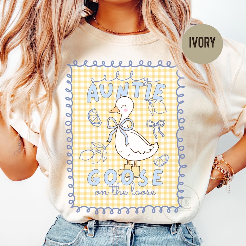 May include: Ivory-colored t-shirt featuring a whimsical design. The graphic includes a cartoon goose with a blue bow, surrounded by lemons and the text "Silly Auntie Goose on the Loose" in blue. The background is a yellow and white gingham pattern.