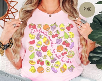 May include: A light pink t-shirt featuring a colorful pattern of various fruits and vegetables, including strawberries, bananas, grapes, and oranges, with the text "Celebrate minds of all kinds" written in a cursive pink font across the center.