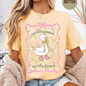 Może przedstawiać: Jasnoż&oacute;łta koszulka z grafiką białej gęsi z kresk&oacute;wki w r&oacute;żowych butach i ż&oacute;łtą kokardą. Tekst "auntie goose on the loose" jest napisany nad i pod gęsią w r&oacute;żowej ramce.