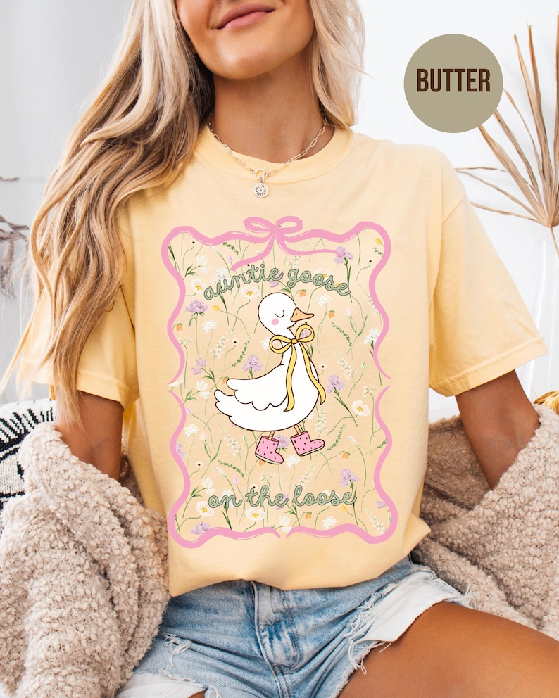 Może przedstawiać: Jasnoż&oacute;łta koszulka z grafiką białej gęsi z kresk&oacute;wki w r&oacute;żowych butach i ż&oacute;łtą kokardą. Tekst "auntie goose on the loose" jest napisany nad i pod gęsią w r&oacute;żowej ramce.
