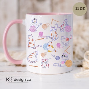 May include: White ceramic mug with a pink handle and rim. The mug features a colorful illustration of cats playing with yarn and knitting needles. The text "11 oz" is printed on the mug.