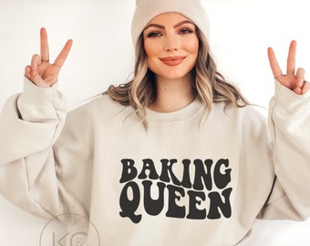 May include: A sand-colored sweatshirt with the text "BAKING QUEEN" in a retro font. The sweatshirt is paired with a matching beanie. The text "For oversized look order 1-2 sizes up" is visible at the top.