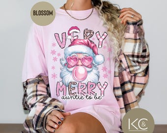 May include: A light pink t-shirt with a graphic of Santa Claus wearing heart-shaped sunglasses and blowing a bubblegum bubble. The text "VERY MERRY auntie to be" is printed below the Santa graphic. The shirt has a long sleeve plaid shirt over it.