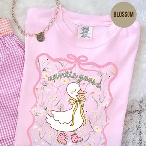 Może przedstawiać: Jasnor&oacute;żowa koszulka z grafiką białej gęsi w ż&oacute;łtej kokardzie i r&oacute;żowych butach, z zielonym napisem "auntie goose". Projekt jest obramowany r&oacute;żową wstążką i kwiatowym wzorem. Widoczny jest r&oacute;wnież srebrny naszyjnik i kraciasta tkanina.