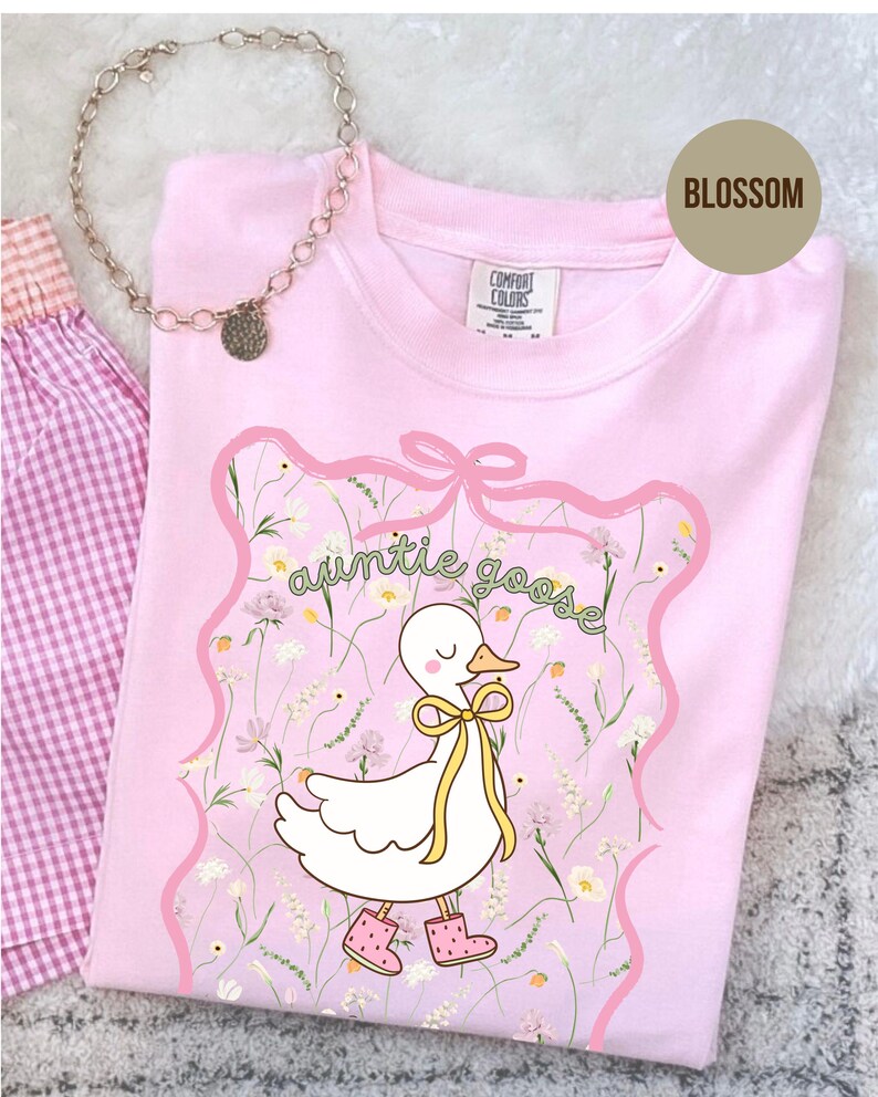 Może przedstawiać: Jasnor&oacute;żowa koszulka z grafiką białej gęsi w ż&oacute;łtej kokardzie i r&oacute;żowych butach, z zielonym napisem "auntie goose". Projekt jest obramowany r&oacute;żową wstążką i kwiatowym wzorem. Widoczny jest r&oacute;wnież srebrny naszyjnik i kraciasta tkanina.