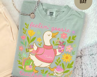 May include: A sage green crewneck t-shirt featuring a graphic of a white goose wearing a pink sweater with the word Teacher printed on it. The goose is surrounded by colorful flowers, a green backpack, a cup, and books with the text feelin' goosie teachin' loosie.