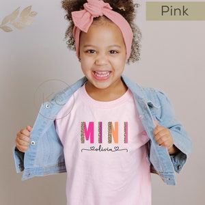 May include: Pink t-shirt with the word "MINI" in colorful letters with a leopard print pattern. The name "Olivia" is written below in a cursive font with hearts on either side.