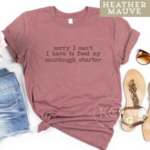 May include: A heather mauve t-shirt with the text "sorry I can't I have to feed my sourdough starter" printed on the front. The shirt is laid flat on a white surface with a pair of sandals and a pair of jeans.