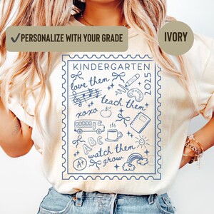 May include: Ivory t-shirt with a blue stamp design, featuring the word "KINDERGARTEN" and school-themed illustrations. The text "love them, teach them, watch them grow" is included. The shirt also has the text "PERSONALIZE WITH YOUR GRADE" and "2025".