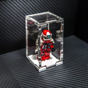 May include: A red and white Lego minifigure with a helmet, displayed in a clear plastic case.